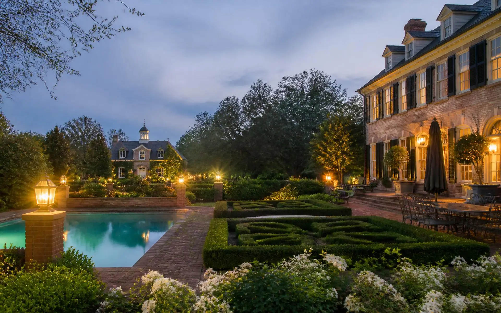 A brick terrace, boxwood parterre garden, pool, and lit guest house behind an Eastover-style estate residence at dusk